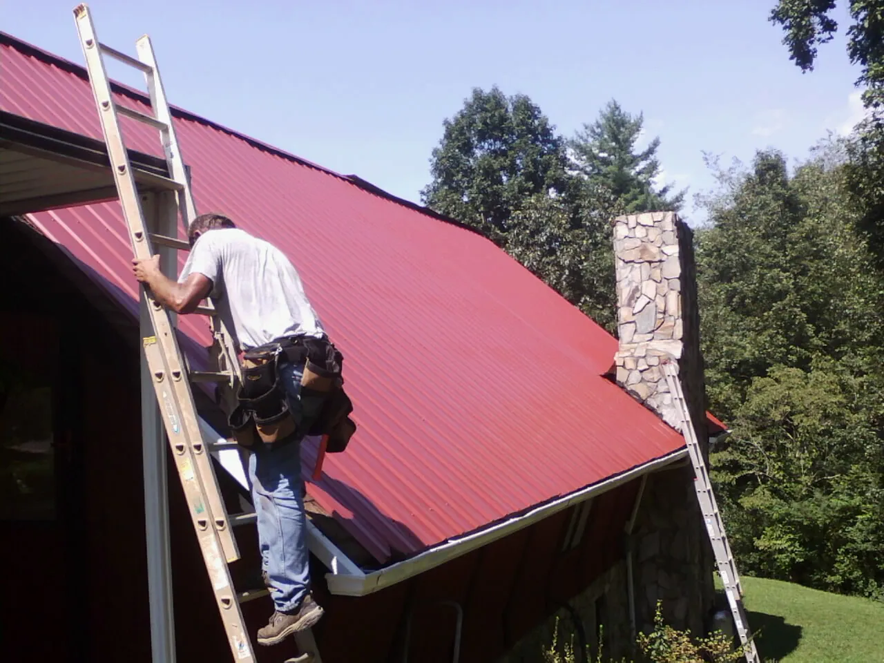 Roofer on a ladder beside a red metal roof and stone chimney — Soffit & Fascia Repair by Roofing Brothers in Firth